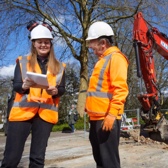 Two employees are assessing work at a construction site with an excavator in the background.