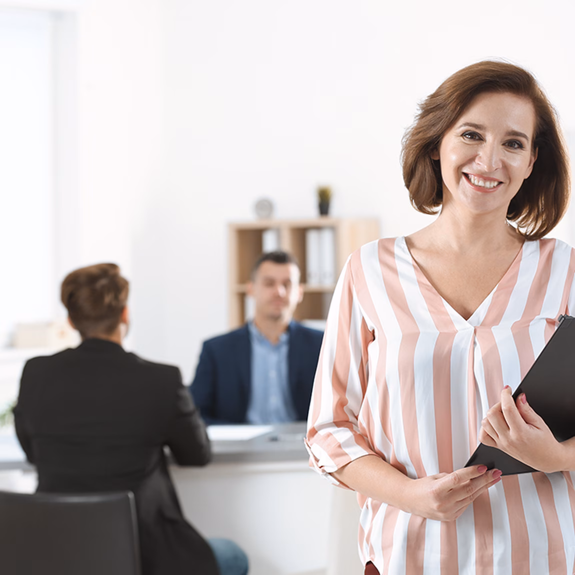 Kantoorprofessional met map in de hand staat in een vergaderruimte terwijl op de achtergrond een gesprek plaatsvindt.