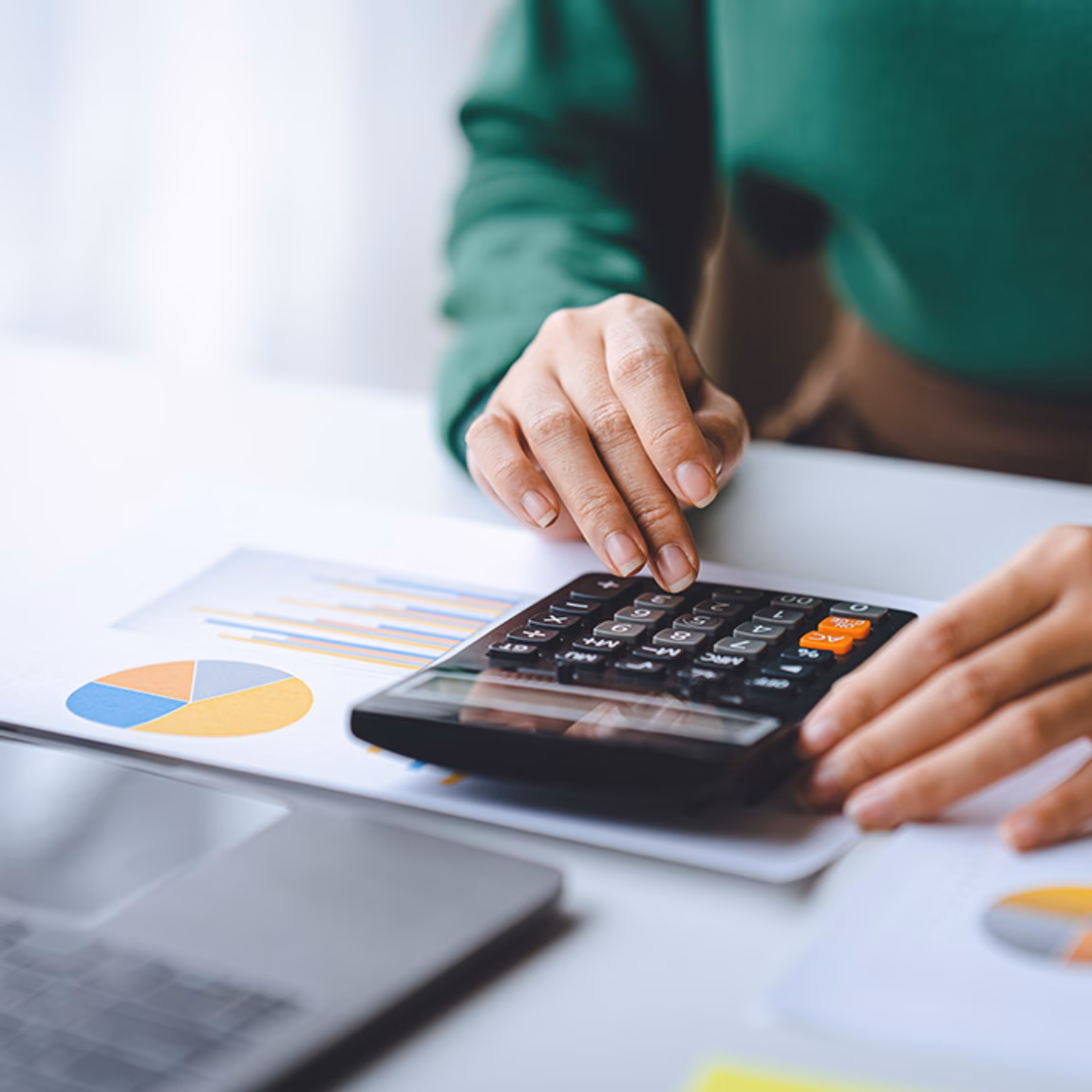 Eine Person nutzt einen Taschenrechner, um Finanzdaten zu analysieren, neben Diagrammen und einem Laptop.