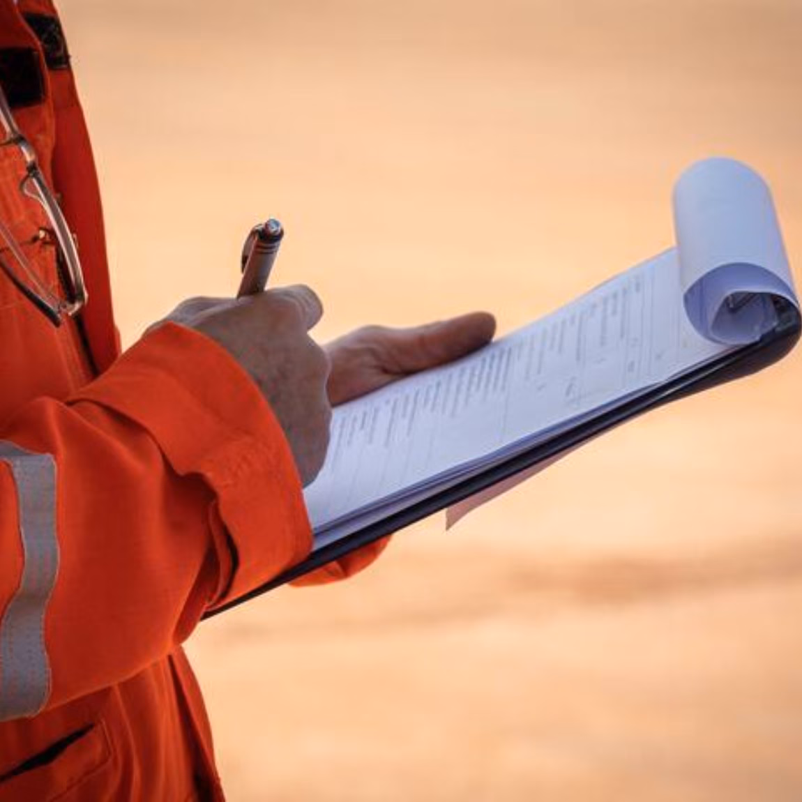 An employee wearing orange safety clothing takes notes in a notebook during an inspection or site visit.