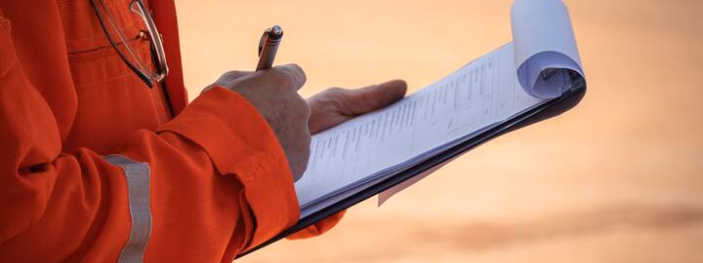 An employee wearing orange safety clothing takes notes in a notebook during an inspection or site visit.