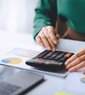 Persoon gebruikt een rekenmachine om financiële gegevens te analyseren naast grafieken en een laptop.