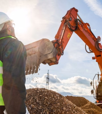 Ein Mitarbeiter in einer Sicherheitsweste überprüft die Arbeiten eines Baggers auf einer Erdbaustelle.