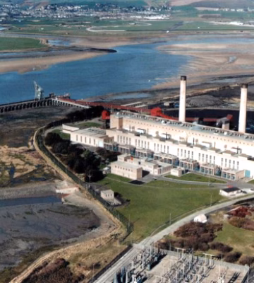Aerial view of an industrial power plant located along a river or in a coastal area, featuring multiple buildings, smokestacks, and loading facilities.