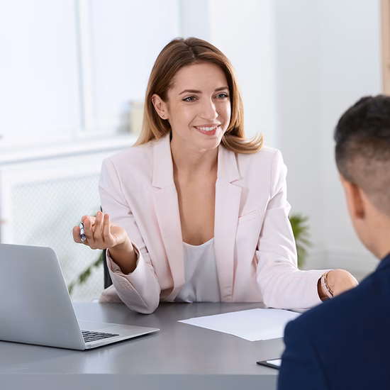 A business meeting between two professionals sitting at a desk with a laptop in an office setting.