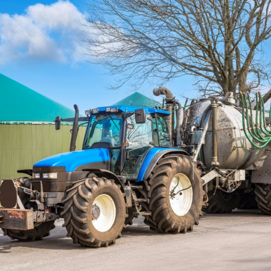 A tractor with a manure tank and a drag-hose spreader is driving down a road next to a fermentation plant.