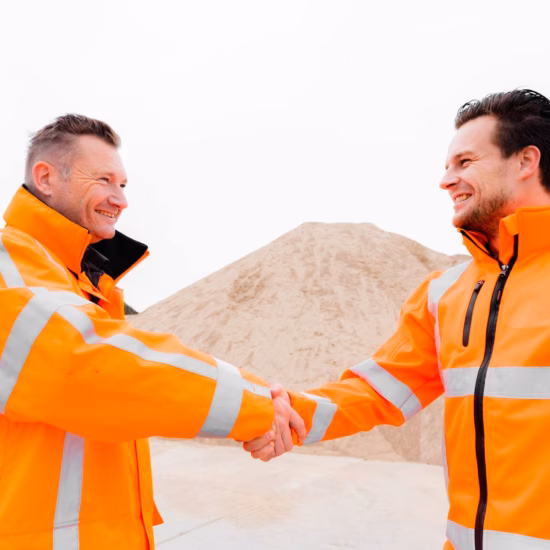 Two employees wearing reflective safety clothing shake hands at an industrial site.