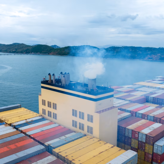 Ein Containerschiff mit Frachtcontainern fährt in der Nähe eines Hafens, während Emissionen aus dem Schornstein sichtbar sind.