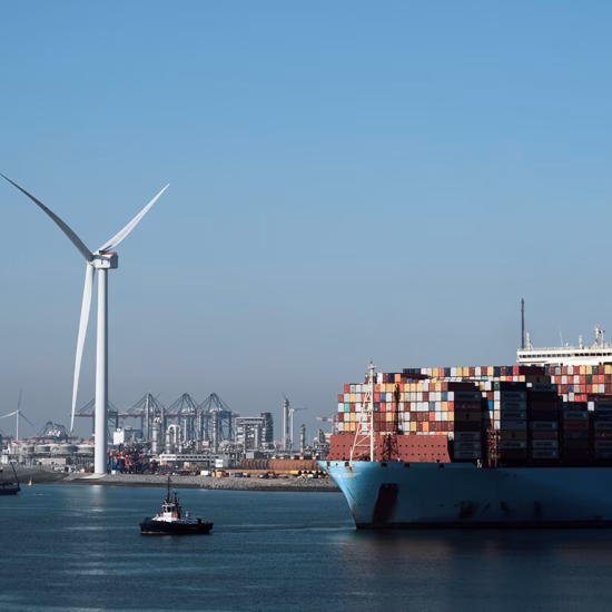 Ein Containerschiff fährt in einem Hafengebiet vor dem Hintergrund einer Windkraftanlage als Symbol für nachhaltige maritime Logistik.