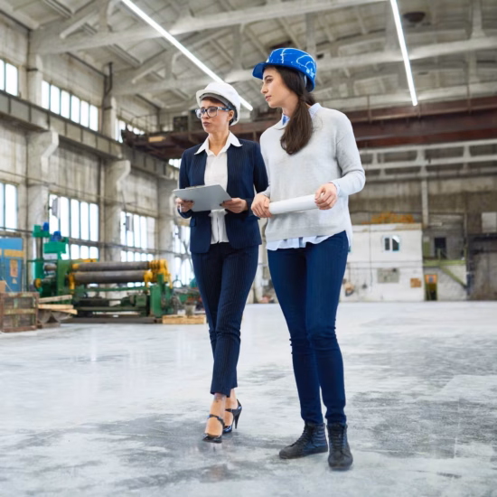 Zwei Mitarbeiter mit Schutzhelmen gehen während einer Inspektion oder eines Standortbesuchs durch eine industrielle Produktionsstätte.