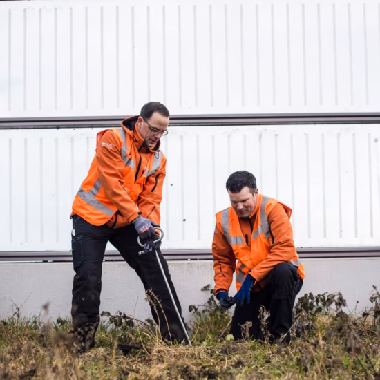 Two employees are conducting inspection and measurement work on the ground near an industrial site.