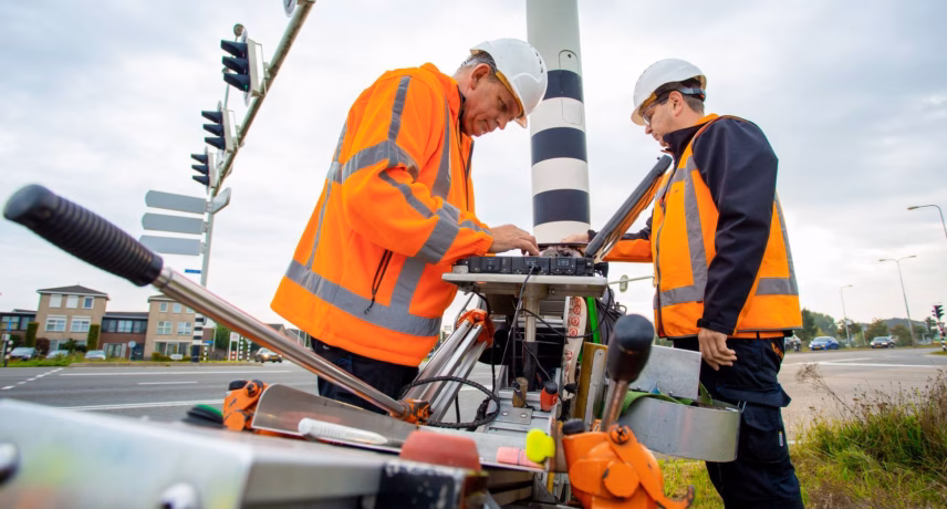 Two employees in safety gear are working on infrastructure along a road using specialized equipment. The image relates to safety, quality management, inspection, and the professional execution of projects in the infrastructure sector.
