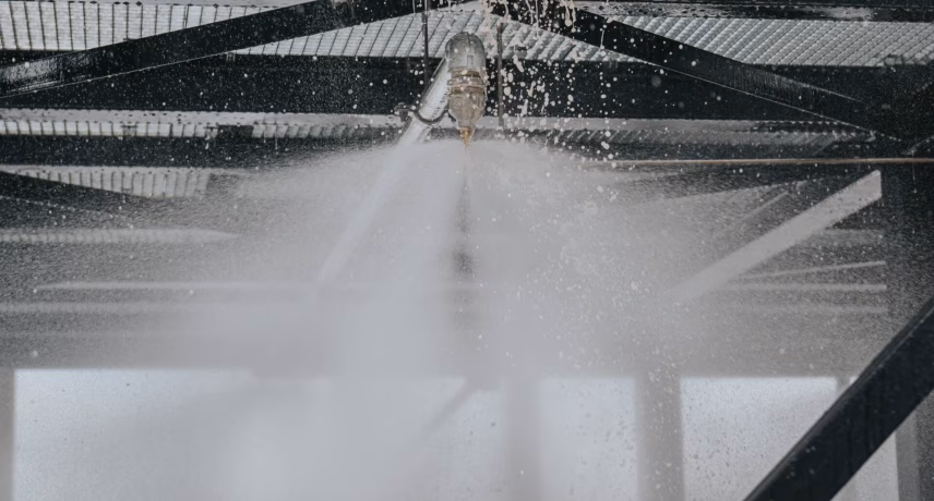 A sprinkler head activates a stream of water from a sprinkler system in an industrial setting. The image relates to fire safety, inspection, system maintenance, and quality assurance.