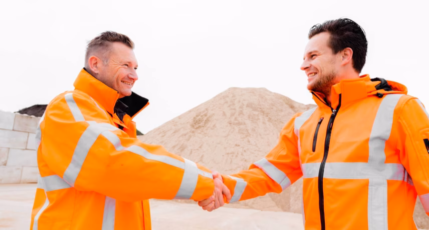 Two employees wearing reflective safety clothing shake hands at an industrial site.