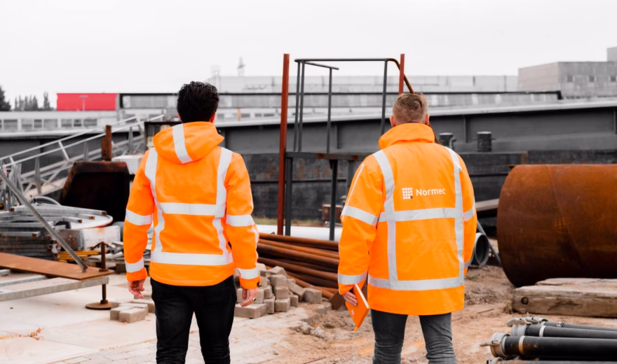 Zwei Mitarbeiter in reflektierender Sicherheitskleidung führen eine Standortbegehung auf einer Industriebaustelle durch.