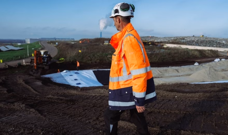 An employee wearing high-visibility safety clothing inspects an outdoor industrial site with earthworks and cordoned-off areas.