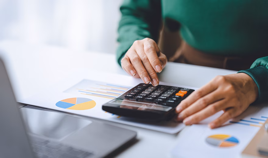 Eine Person nutzt einen Taschenrechner, um Finanzdaten zu analysieren, neben Diagrammen und einem Laptop.