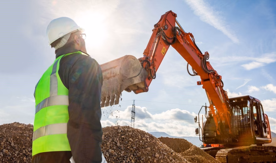 An employee wearing a safety vest inspects the work of an excavator at an earthmoving site.