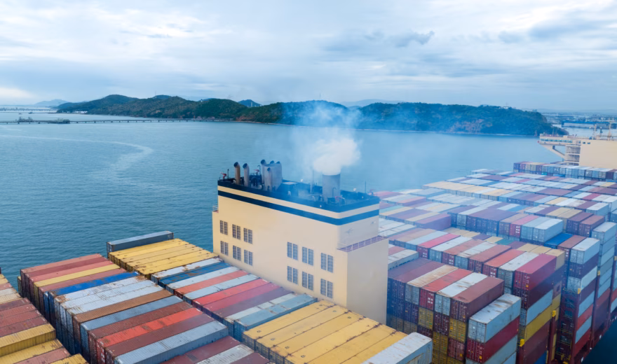 A container ship carrying cargo containers is sailing near a port while emissions are visible from its smokestack.