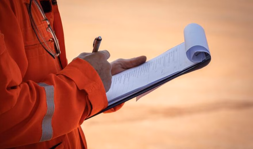 An employee wearing orange safety clothing takes notes in a notebook during an inspection or site visit.