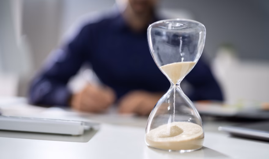 Close-up Of Hourglass In Front Of Businessperson's Hand Calculating Invoice