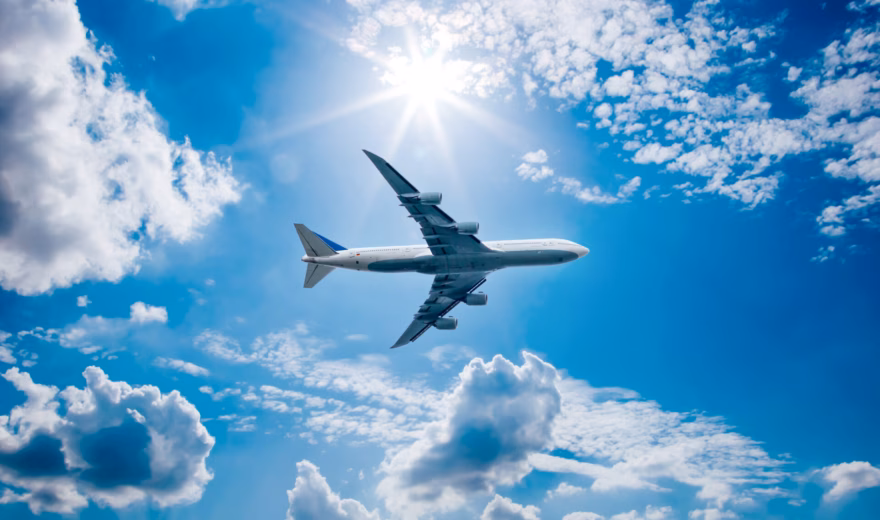 A passenger plane is flying at cruising altitude beneath a clear sky with clouds.