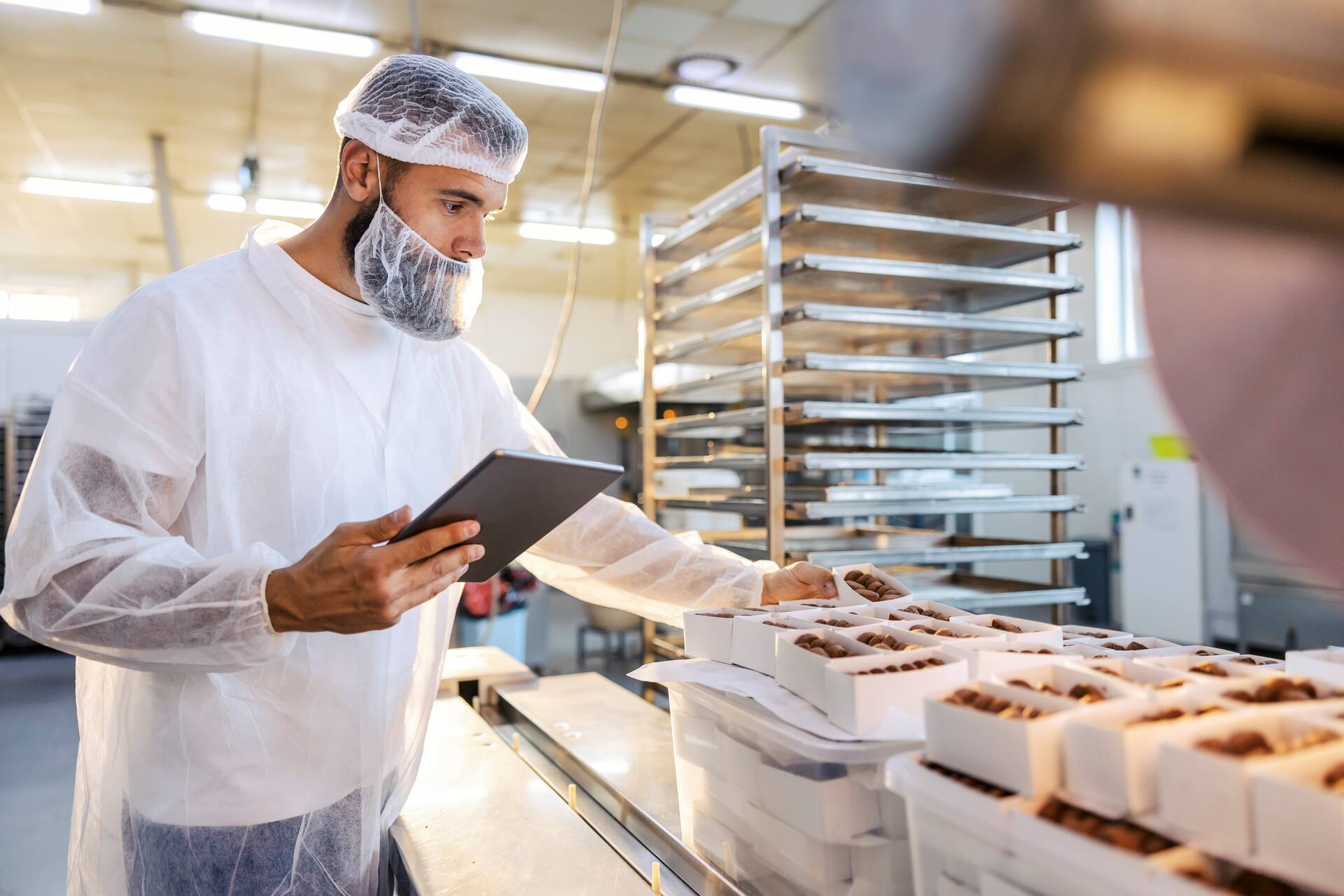 A food inspector in a sterile white uniform is holding the tablet and looking at collected cookies.