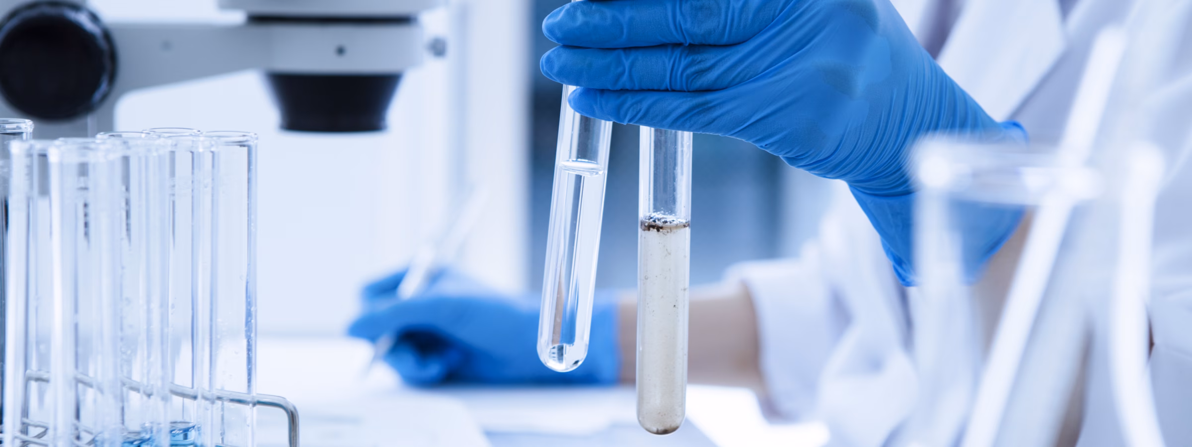 A laboratory researcher holds test tubes containing samples next to a microscope for analysis.