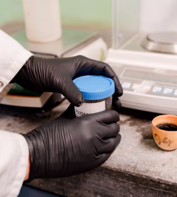 Laboratoriummedewerker met zwarte handschoenen houdt een monsterpot vast naast een digitale laboratoriumweegschaal.