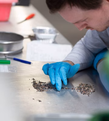 Laboratoriummedewerker met blauwe handschoenen onderzoekt kleine materiaaldeeltjes op een werktafel.