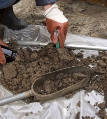 Medewerker verzamelt een grondmonster met een metalen schep en onderzoeksapparatuur voor bodemonderzoek.