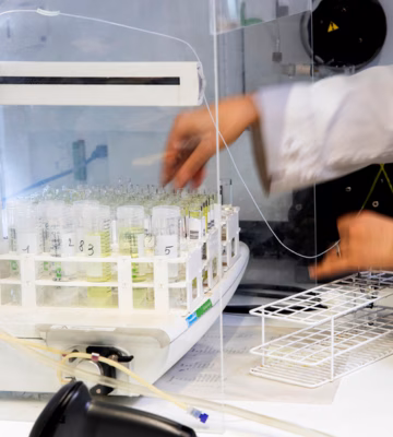 In een laboratorium plaatst een medewerker een rek met reageerbuizen in een geavanceerd analyseapparaat. De afbeelding toont werkzaamheden rondom laboratoriumonderzoek, monsteranalyse en kwaliteitscontrole binnen een onderzoeks- of testomgeving.