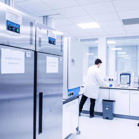Een helder verlicht laboratorium met roestvrijstalen koel- en opslagapparatuur langs de wand. Op de achtergrond werkt een laboratoriummedewerker aan een werkstation voor onderzoek, monsterverwerking of kwaliteitscontrole. De afbeelding benadrukt een gecontroleerde en hygiënische laboratoriumomgeving.