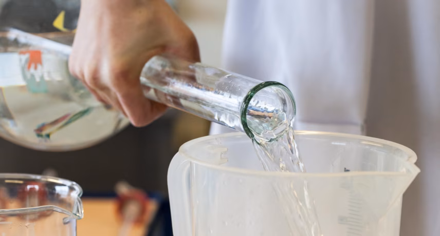 A person pours a clear liquid from a glass bottle into a transparent measuring cup, suitable for laboratory testing, quality control, and analysis.
