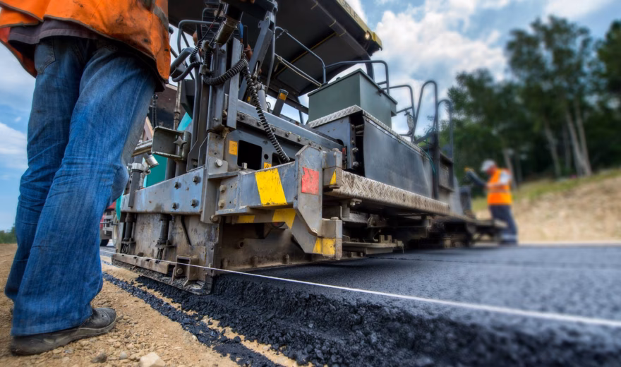 Asfalteermachine legt nieuw asfalt aan op een weg terwijl medewerkers in veiligheidskleding toezicht houden op de werkzaamheden.