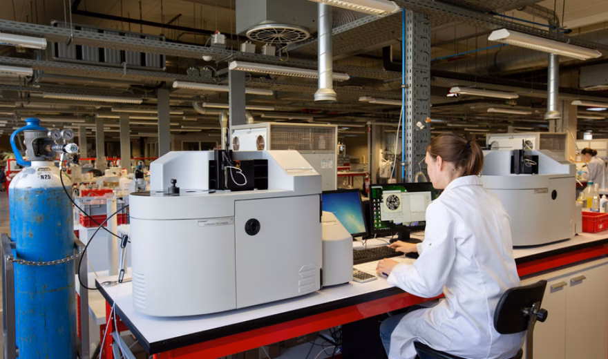 Laboratoriummedewerker in een witte labjas werkt achter een computer naast geavanceerde analyseapparatuur in een laboratorium.