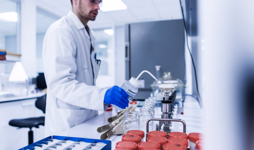 Laboratoriummedewerker in een witte labjas en blauwe handschoenen werkt met monsterbuizen en analyseapparatuur in een laboratorium.