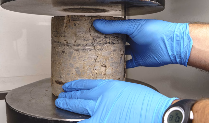 Laboratoriummedewerker met blauwe handschoenen plaatst een cilindervormig betonmonster in een druktestmachine.