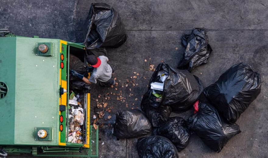 A waste collection vehicle surrounded by trash bags during waste processing on a paved area.