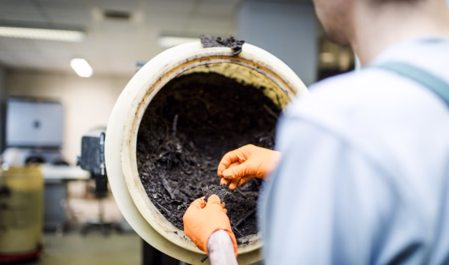A person examines a dark organic sample in a rotating laboratory drum.