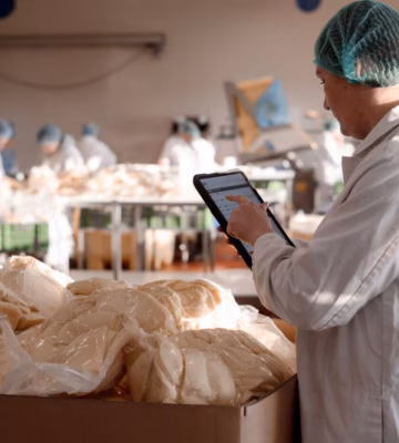 Persoon in beschermende werkkleding gebruikt een tablet voor digitale registratie of kwaliteitscontrole bij voedingsproducten in een productieomgeving.