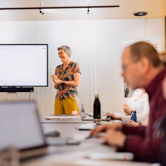 The trainer presents during a business workshop while participants work on their laptops at a conference table.