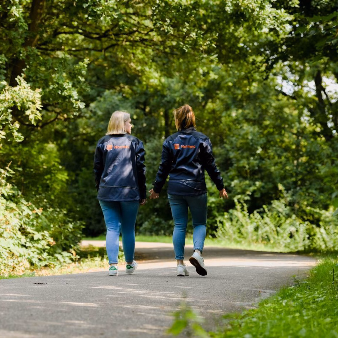Twee personen wandelen over een verhard pad door een groene, bosrijke omgeving.