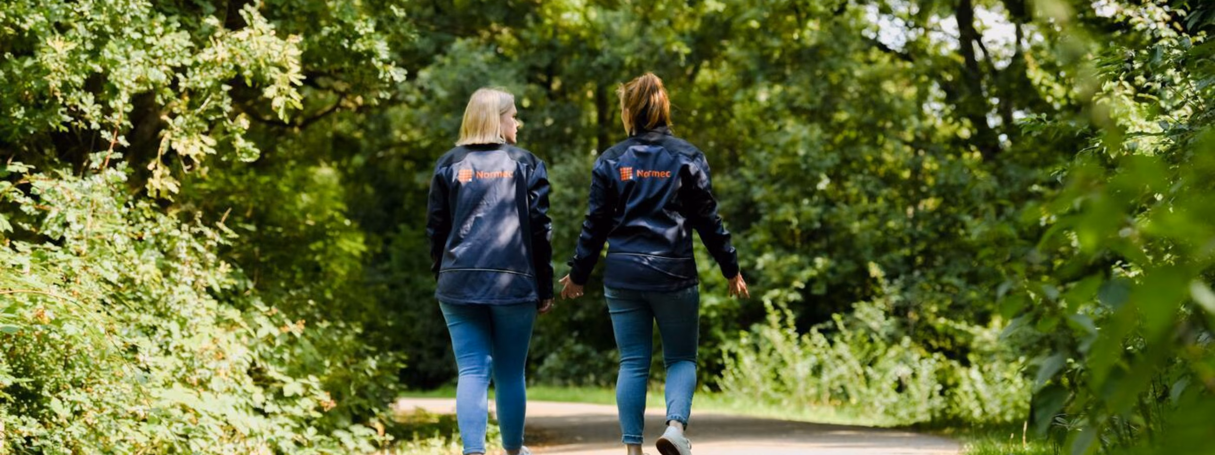 Twee personen wandelen over een verhard pad door een groene, bosrijke omgeving.