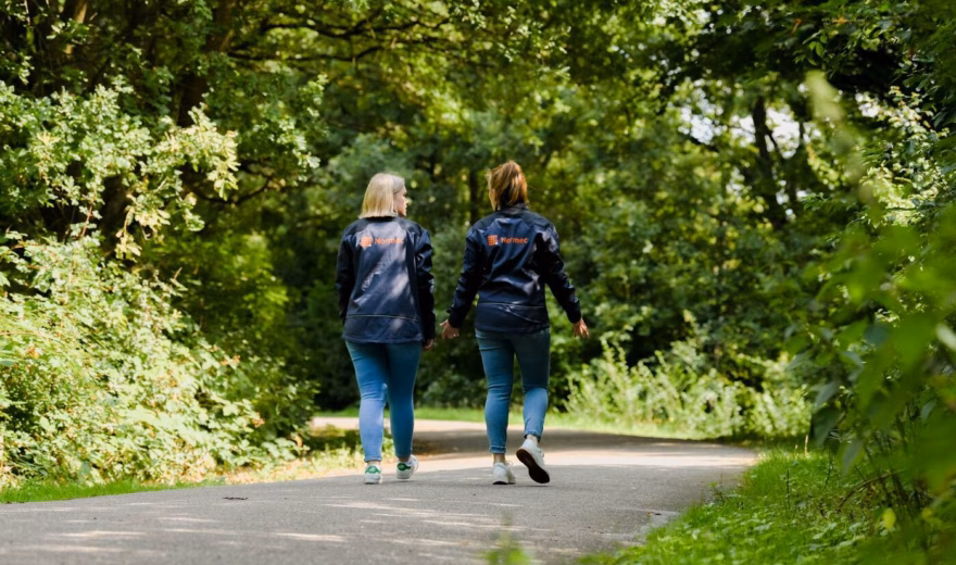 Twee personen wandelen over een verhard pad door een groene, bosrijke omgeving.