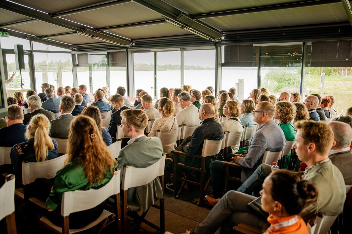 Publiek volgt een presentatie of kennissessie tijdens een bijeenkomst in een conferentie- of trainingsruimte.
