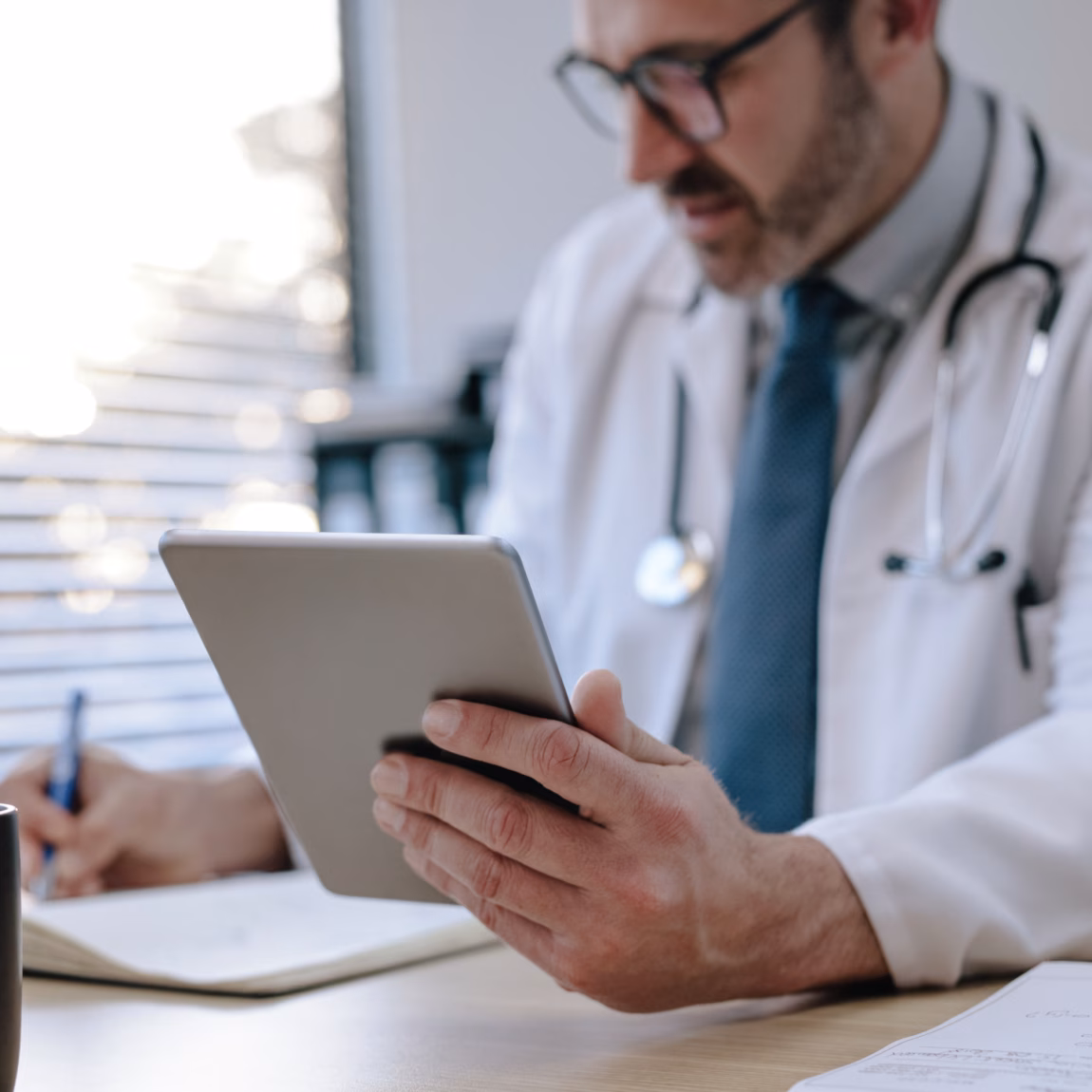A healthcare professional wearing a stethoscope uses a tablet and takes notes at a desk in a medical setting.
