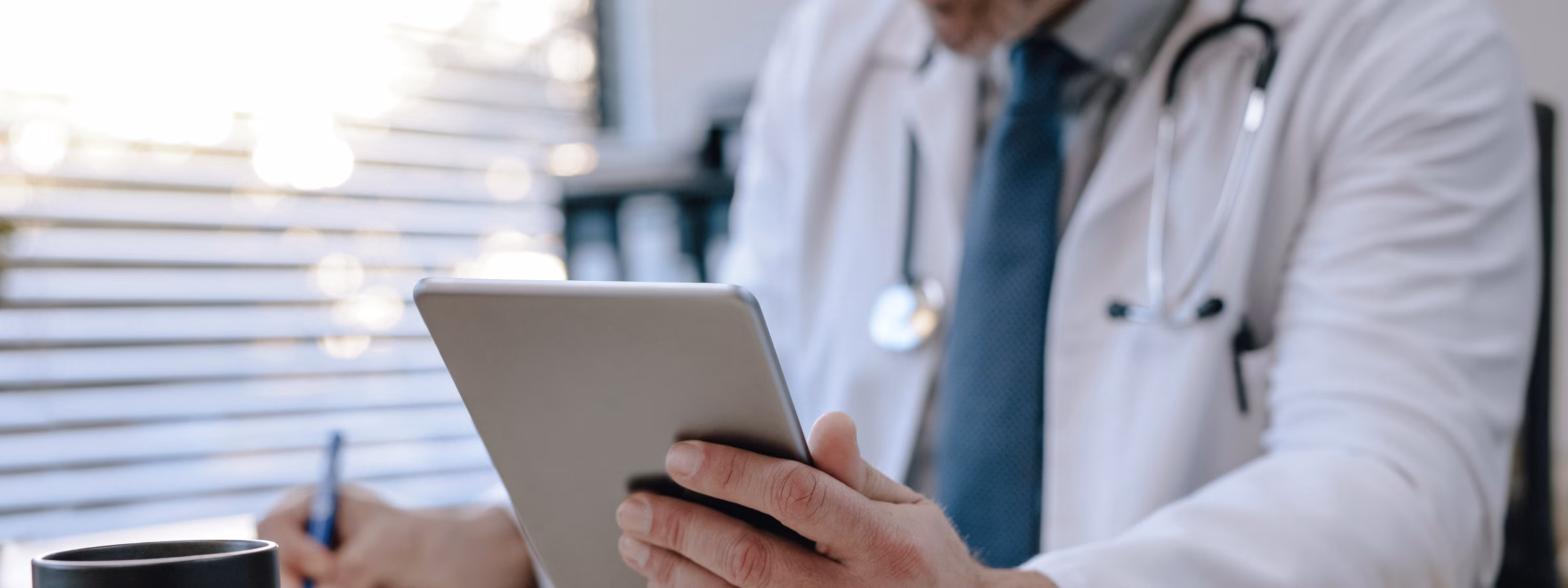 A healthcare professional wearing a stethoscope uses a tablet and takes notes at a desk in a medical setting.