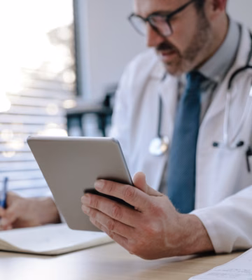 Zorgprofessional met stethoscoop gebruikt een tablet en maakt aantekeningen aan een bureau in een medische werkomgeving.