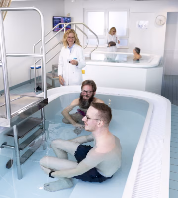 Hydrotherapiebecken mit drei Personen im Wasser und einer Pflegekraft neben dem Becken in einer Rehabilitationsumgebung.